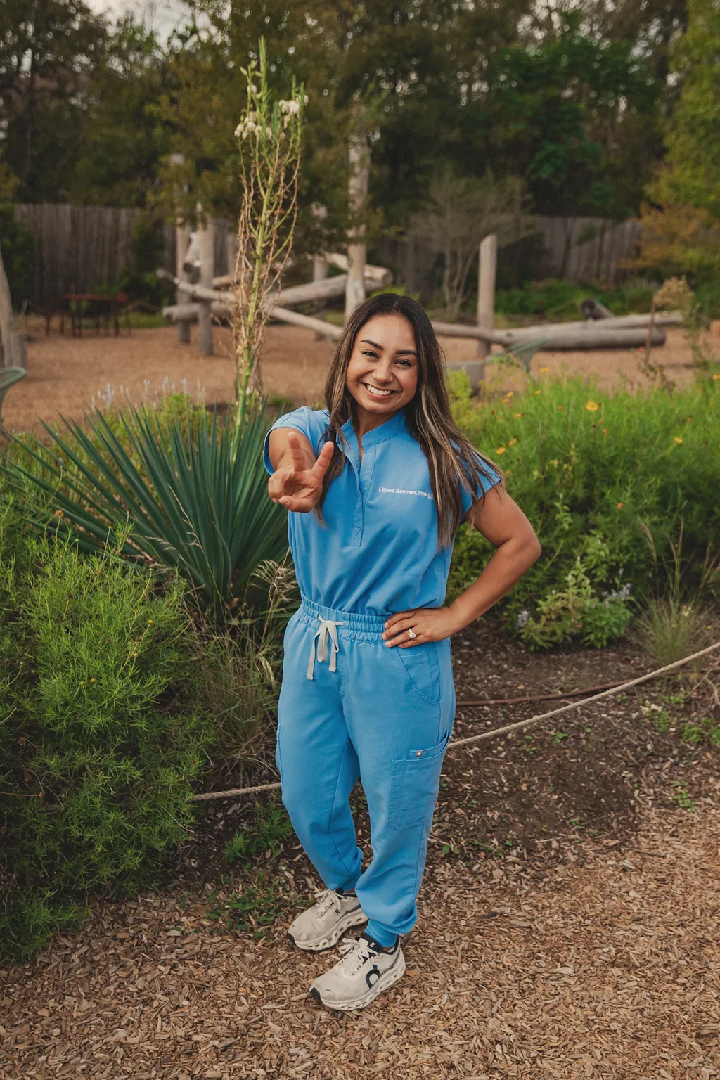 Liliana Damron, APRN, FNP-BC, smiling in light blue scrubs embroidered with her name, in a native-plant garden.