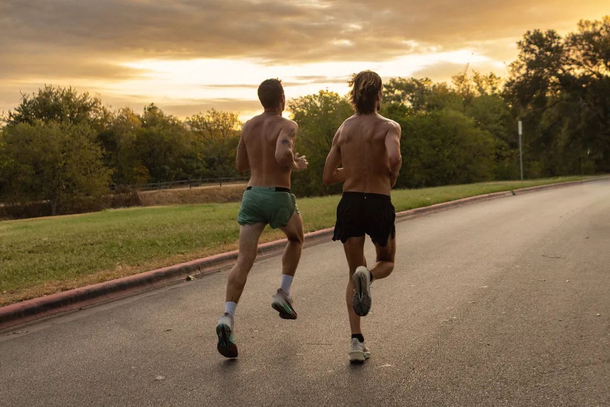 Two shirtless runners heading down a park road under a golden sunset sky.