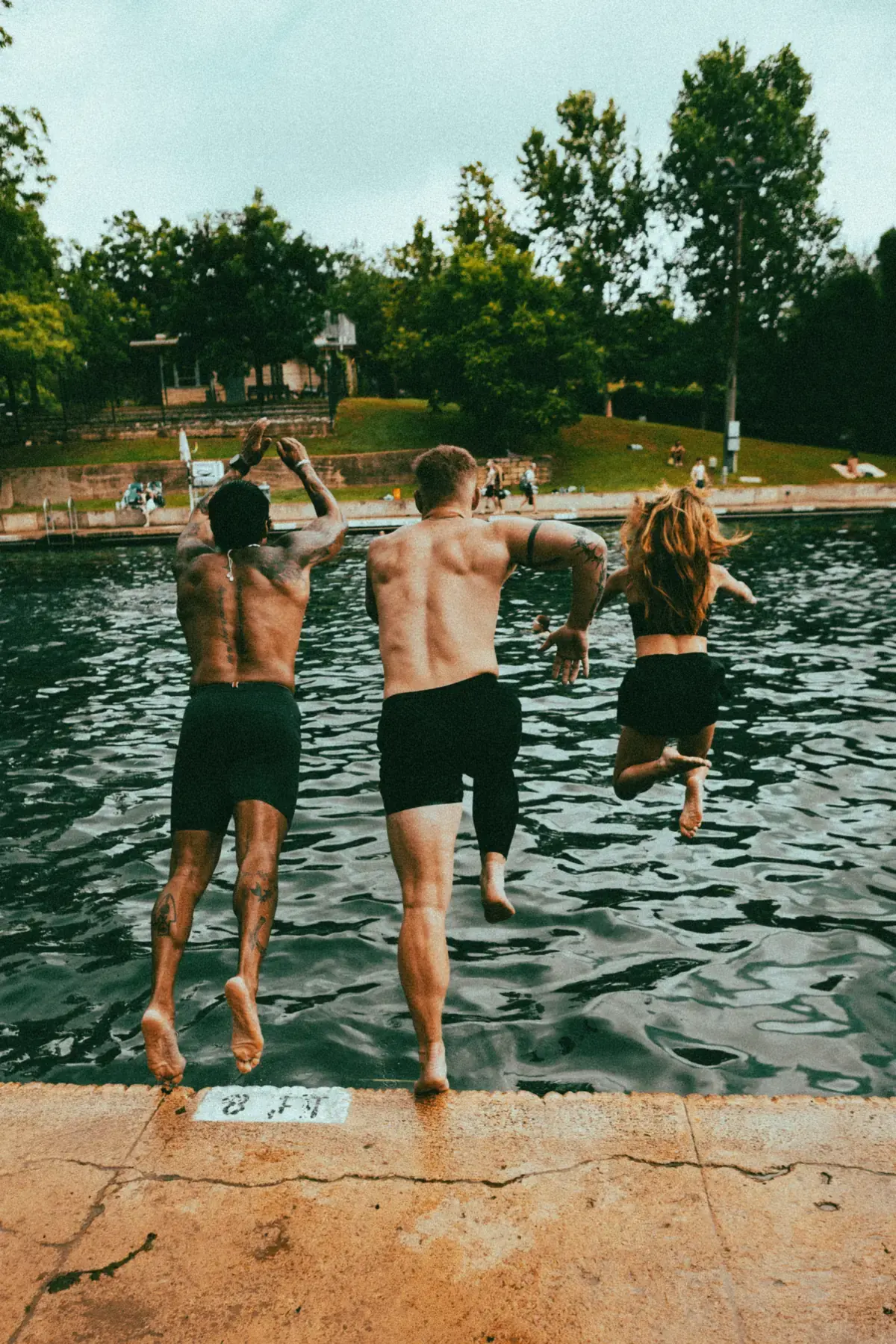 Three friends jumping off the edge of Barton Springs Pool by the 8 FT depth marker, seen from behind.