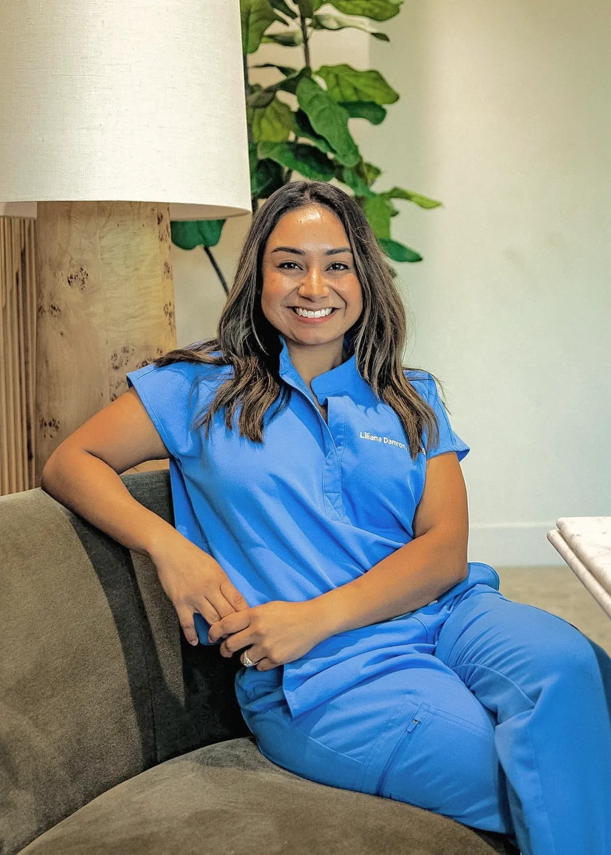 Liliana Damron, APRN, FNP-BC, smiling while seated on a couch in light blue scrubs.